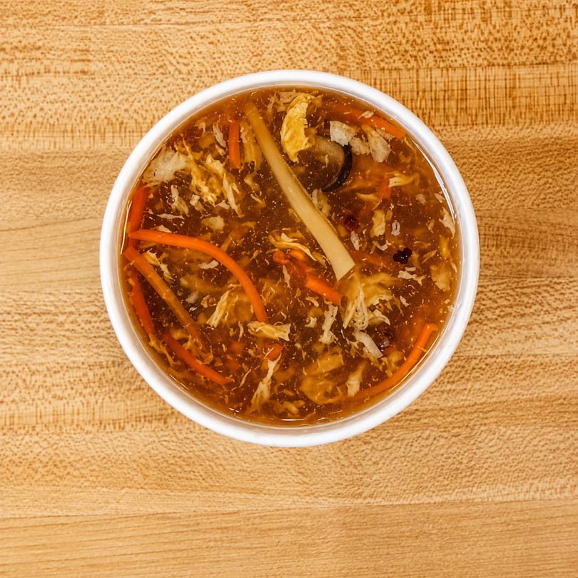Overhead view of a bowl of hot and sour soup with carrots, mushrooms, and egg on a wooden table.