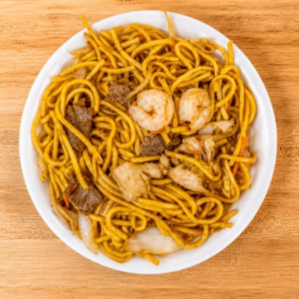 A white bowl of lo mein noodles with shrimp, beef, chicken, onions, and carrots on a wooden table.